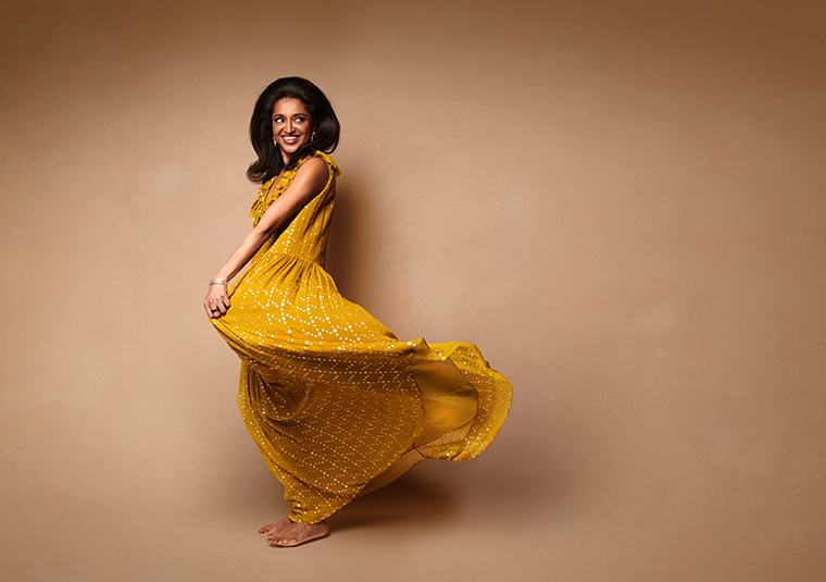 Sindhu Vee in a flowy yellow dress against a brown background.