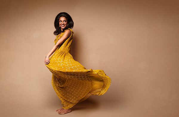 Sindhu Vee in a flowy yellow dress against a brown background.