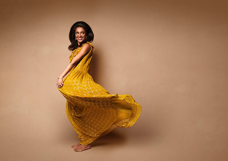 Sindhu Vee in a flowy yellow dress against a brown background.