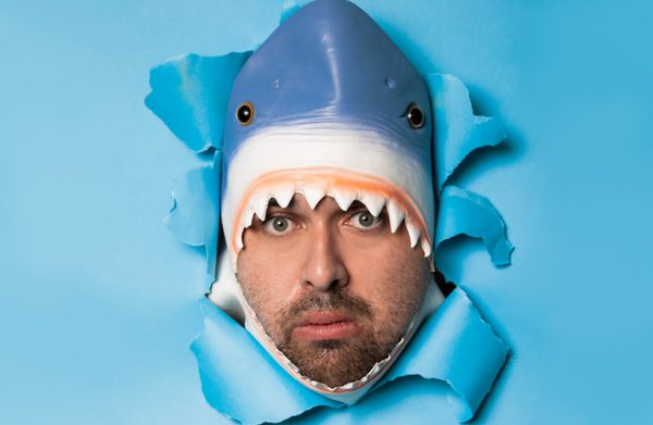 An actor in a shark's head bursts through a sheet of blue paper, their face pokes through the shark's mouth