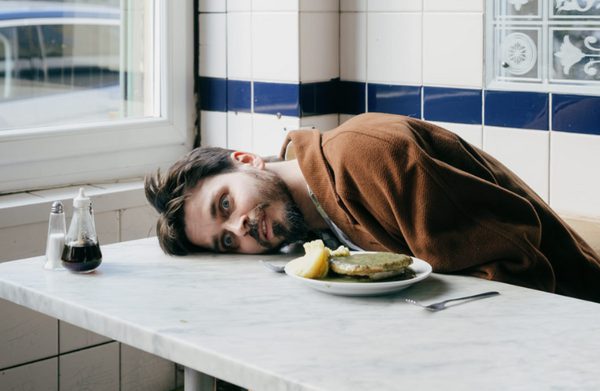 Joz Norris sitting at a restaurant table, head laying on the surface of the table with a wide eyed expression on his face
