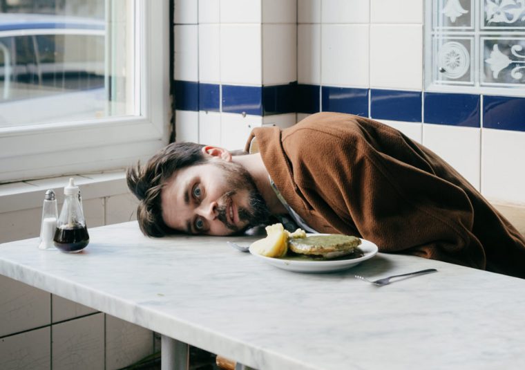 Joz Norris sitting at a restaurant table, head laying on the surface of the table with a wide eyed expression on his face