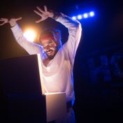 Joz Norris in a white track suit with arms raised, looking looking menacingly at a box with a stark light on his face.