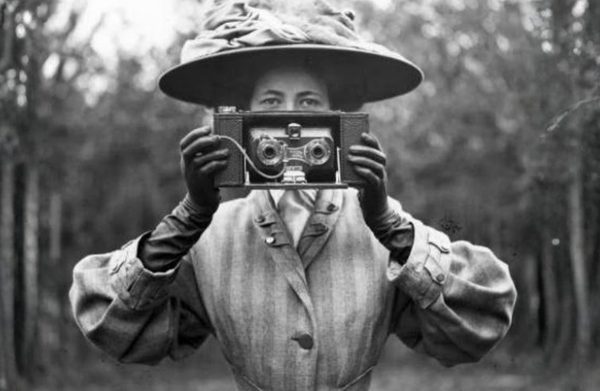 In a Victorian style photograph, portraying Rosalie Chichester, Helena Payne holds up a period-accurate camera. She looks out at us over the camera.
