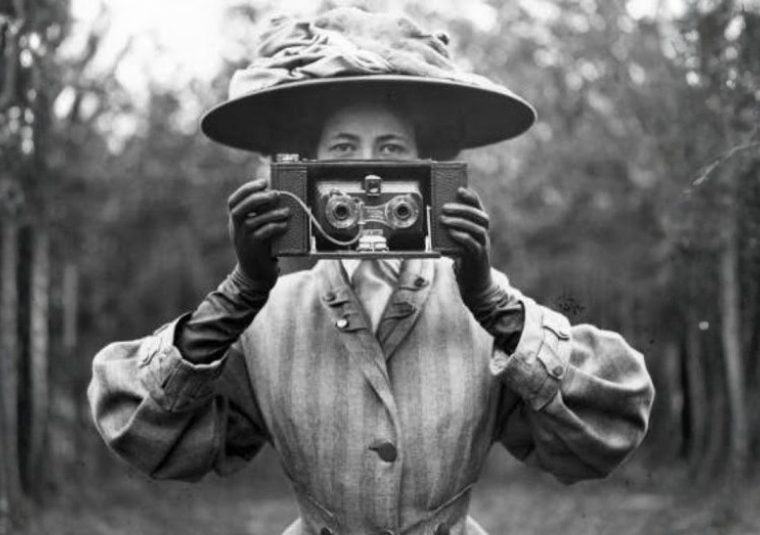In a Victorian style photograph, portraying Rosalie Chichester, Helena Payne holds up a period-accurate camera. She looks out at us over the camera.