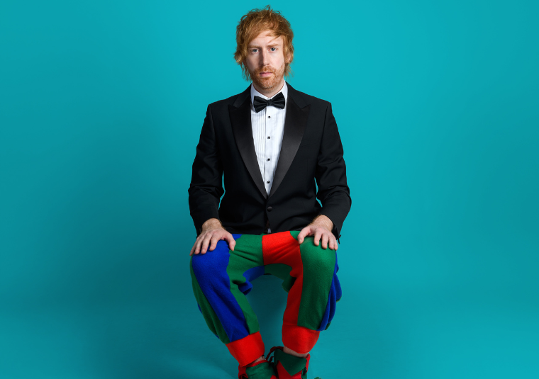 Comedian Mark Simmons sat against a bright blue background wearing a suit and bowtie on top and a jester for trousers and shoes.