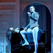 Titania asleep on a piano as Oberon holds a flower and looks towards the crowd.