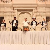 Cast of A Midsummer Night's Dream during a feast scene with actors dressed as servants standing behind a long covered table.