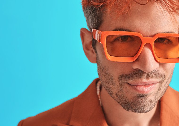 Ivo Graham in bright orange sun glasses and suit against a bright blue background.