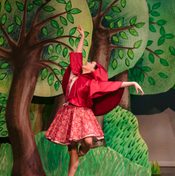 Julie Nunes in Little Red Riding Hood dressed in a pink dress, red cape, bright red ballet shoes in front of a whimsical forest backdrop.