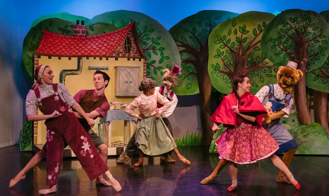 All cast members dressed in whimsical costumes dancing in pairs in front of a whimsical cottage backdrop in a forest.