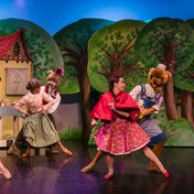 All cast members dressed in whimsical costumes dancing in pairs in front of a whimsical cottage backdrop in a forest.
