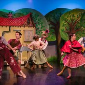 All cast members dressed in whimsical costumes dancing in pairs in front of a whimsical cottage backdrop in a forest.