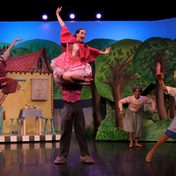 All the cast dancing in pairs against a whimsical forest and cottage backdrop.
