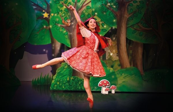 Julie Nunes in Little Red Riding Hood in a pink dress, red cape, red ballet shoes in front of a whimsical painted forest scene.
