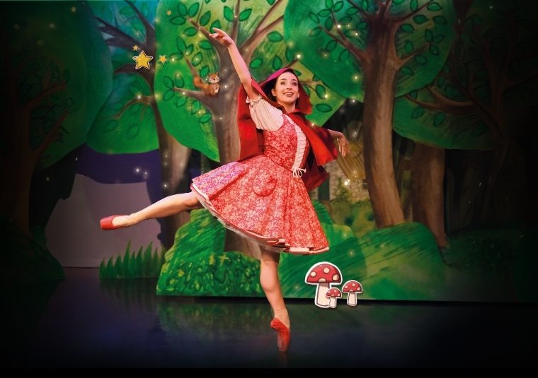 Julie Nunes in Little Red Riding Hood in a pink dress, red cape, red ballet shoes in front of a whimsical painted forest scene.