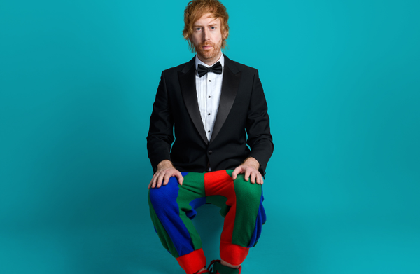 Comedian Mark Simmons sat against a bright blue background wearing a suit and bowtie on top and a jester for trousers and shoes.