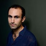 Actor and activist Khalid Abdalla in a blue button up top against a dark grey backdrop