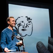 Khalid Abdalla on stage smiling as he holds a drawing of his that is projected in real time behind him.