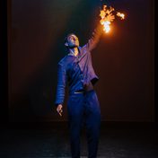 Khalid Abdalla on stage with one arm raised as fire engulfs his left hand, his expression calm.