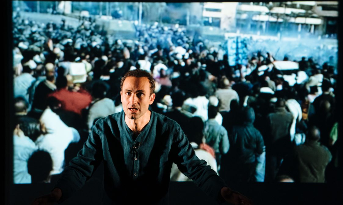 Khalid Abdalla on stage with a projection of a crowd behind him.