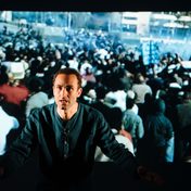 Khalid Abdalla on stage with a projection of a crowd behind him.