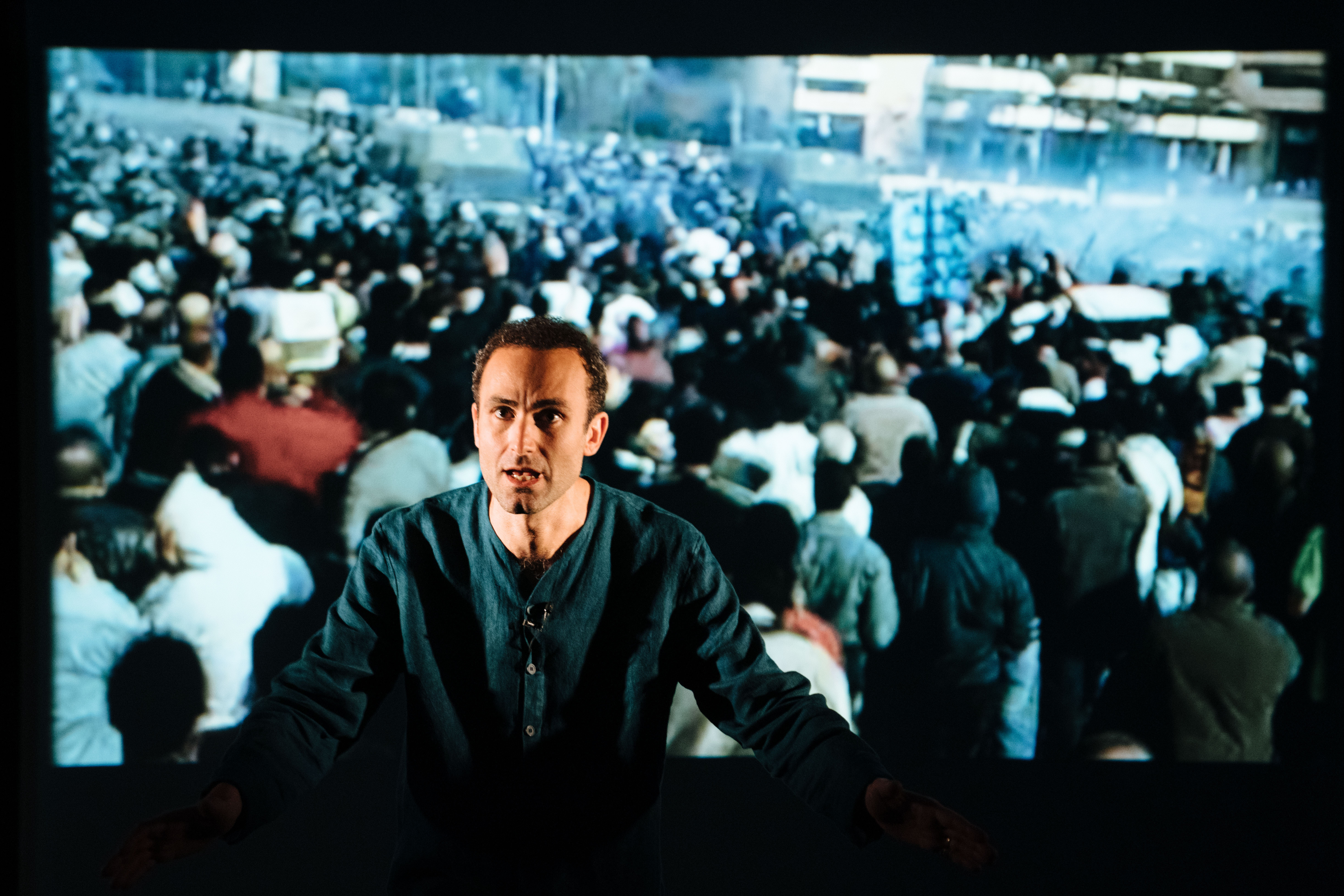 Khalid Abdalla on stage with a projection of a crowd behind him.