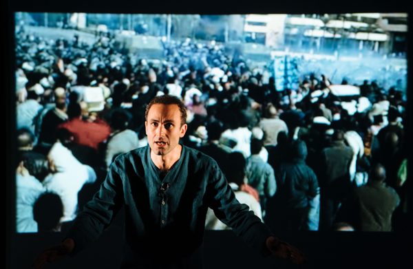 Khalid Abdalla on stage with a projection of a crowd behind him.