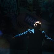 Khalid Abdalla on stage with dramatic lighting as projections of protests appear behind him.