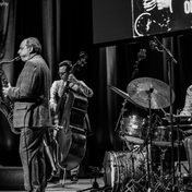 Black and white photo of the band paying on stage with musicians playing a saxophone, drums, and cello.