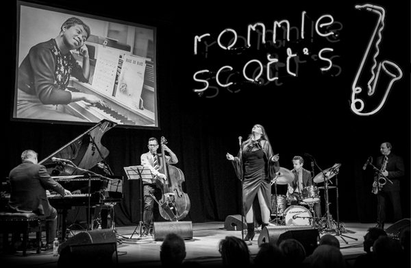 Black and white image of a previous gig of Ronnie Scott's Jazz Club on Tour with the band at centre stage and neon lights in the background with a velvet backdrop.