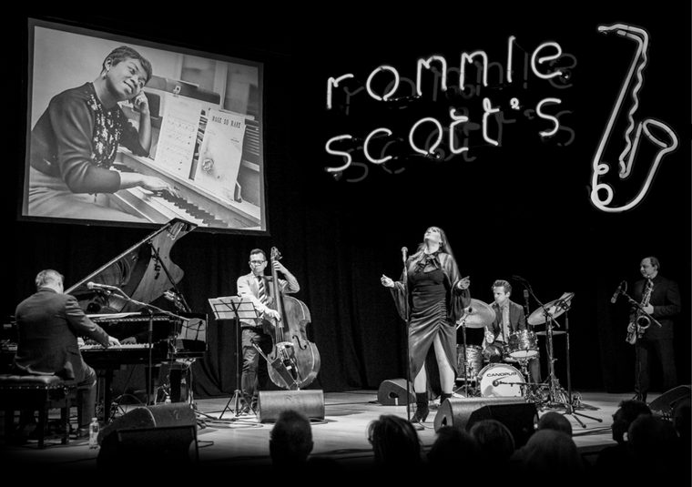 Black and white image of a previous gig of Ronnie Scott's Jazz Club on Tour with the band at centre stage and neon lights in the background with a velvet backdrop.