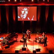 The band on stage with the singer and musicians playing the piano, drums and cello.