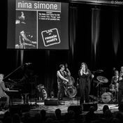 Black and white photo of the band on stage with a singer and musicians playing the piano, drums and cello.