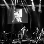 Black and white photo of the band on stage with a singer and musicians playing the piano, drums and cello.