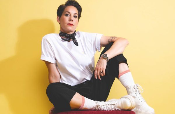 Suzi Ruffell in a white top, black scarf and black trousers sitting against a yellow wall.