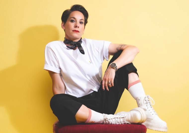 Suzi Ruffell in a white top, black scarf and black trousers sitting against a yellow wall.
