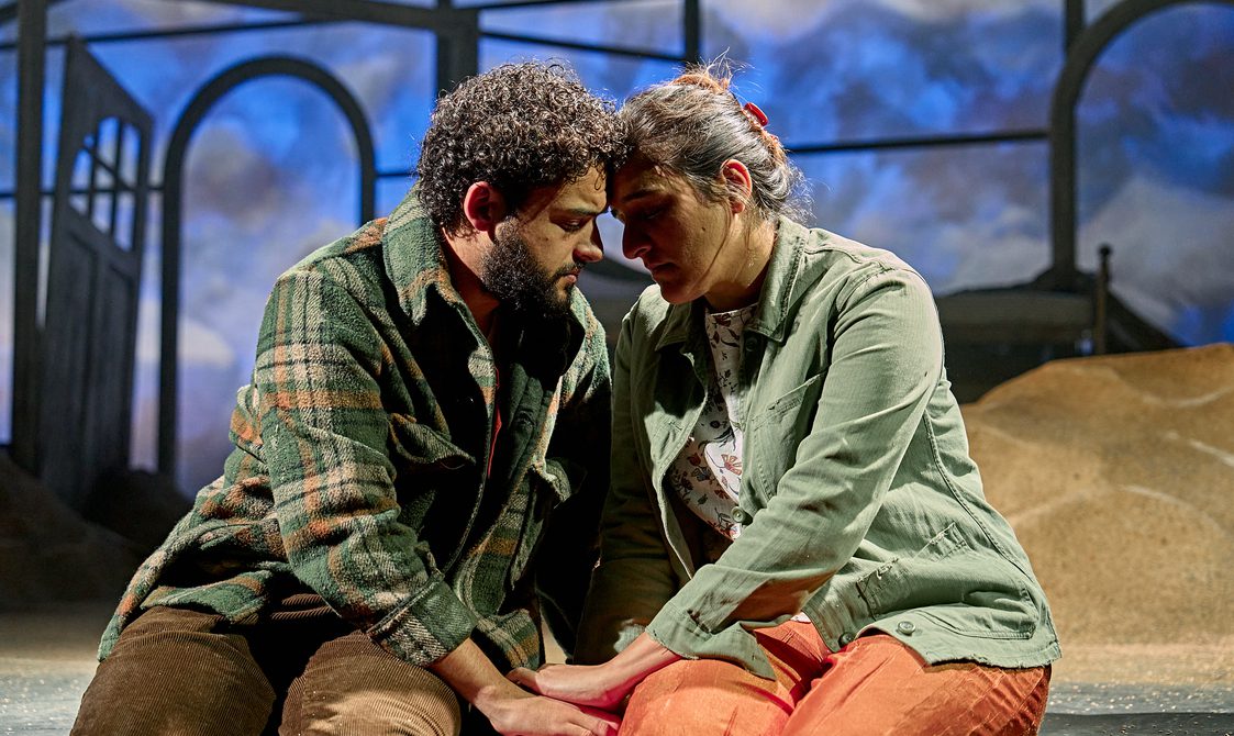 A man and woman sit together at the front of the stage with their eyes closed, holding hands, and heads touching in an intimate, emotional scene.
