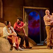 A man sits with an older woman and gentleman in conversation as the man and gentleman point up stage.