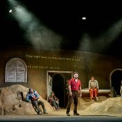 The cast of The Beekeeper of Aleppo in a wide angle shot of the stage including a backdrop of an simple interior of a house, with mounds of dirt or sand which most characters are sitting in with a man at the centre looking towards the crowd.
