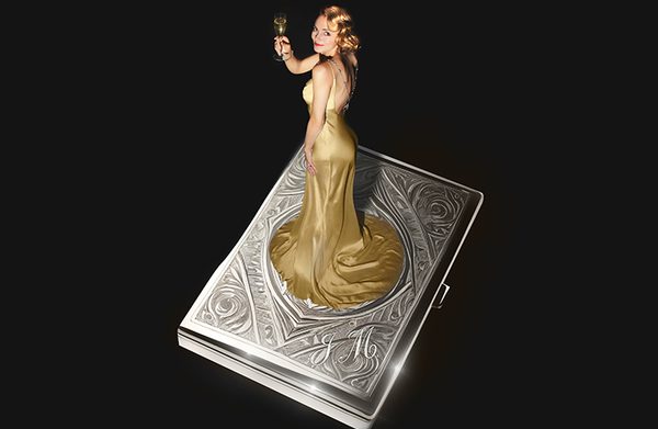 A woman in a vintage gold dress and bright gold hair holding up a flute of champagne standing on top of a vintage cigarette case with a floral pattern embossed on the top and the initials J M.