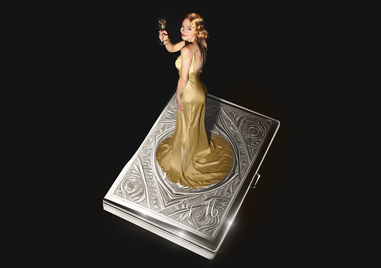 A woman in a vintage gold dress and bright gold hair holding up a flute of champagne standing on top of a vintage cigarette case with a floral pattern embossed on the top and the initials J M.