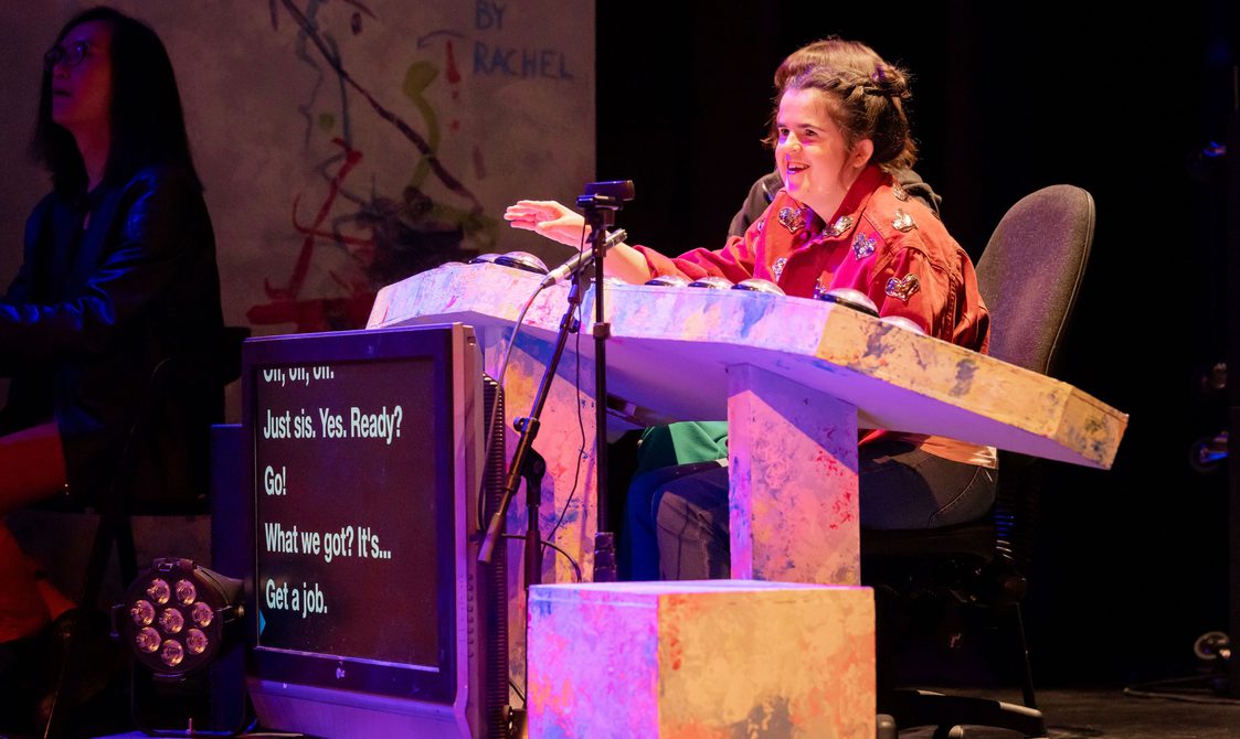 Rachel in a pink jacket sat at her desk with buttons and a captions screen.