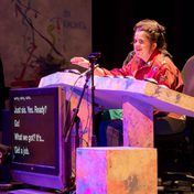 Rachel in a pink jacket sat at her desk with buttons and a captions screen.