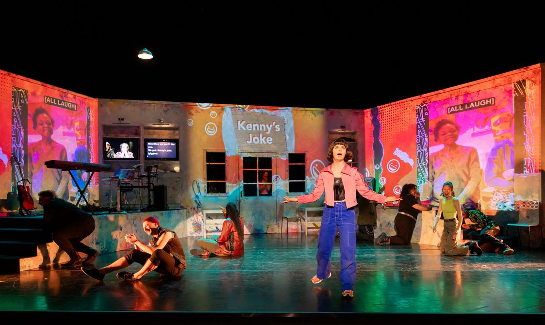 A wide shot of the stage with an actor out in front singing as other actors are on the ground laughing around her. Large projections of people laughing cover the three walls of the set.