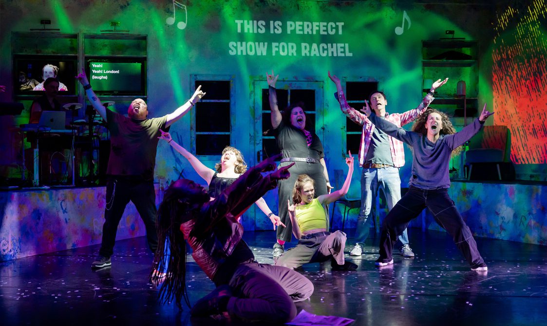A group of actors on stage all with their hands raised as they sing at the top of their lungs with the projection 'This is Perfect Show for Rachel' behind them.