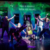 A group of actors on stage all with their hands raised as they sing at the top of their lungs with the projection 'This is Perfect Show for Rachel' behind them.