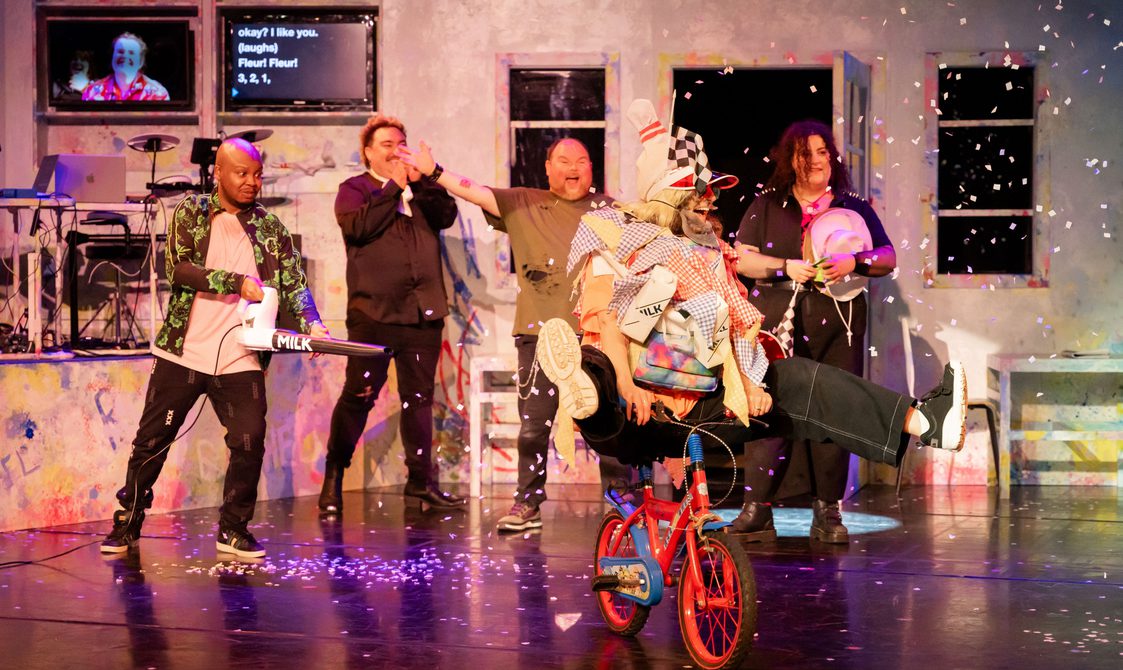 A goofy scene of actors throwing confetti at a central figure who is dressed up and riding a miniature bicycle around stage.b