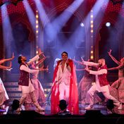A major dance scene with an actor out in front in a striking white suit and red cape with backup dancers posing around him.