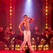 An actor smising on stage while striking a dramatic pose with photographers behind him swooning.